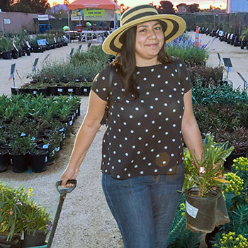 Woman poses for camera, holding two small container plants in one hand and a wagon full of potted plants in the other. The background features the scene of the Plant Sale at the Springs Preserve.