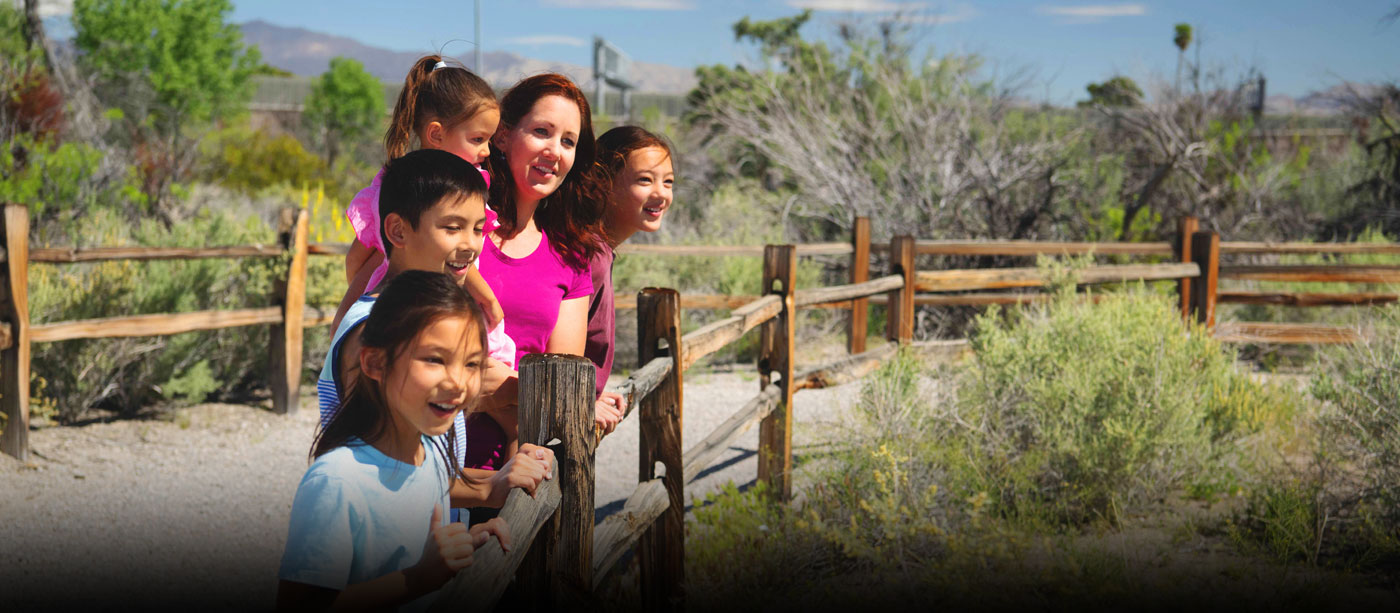 Female adult and four children stand at wooden fence and gaze into distance with the Springs Preserve trails and desert shrubbery in the background.