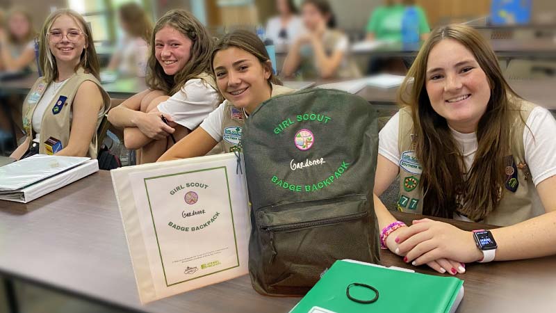 Four Girl Scouts from Troop 777 smile at the camera while seated in a classroom at the Springs Preserve. On the desk in front of them is a black backpack with the words "Girl Scout Gardener Badge Backpack" embroidered on it, as well as several binders and folders related to the Badge Backpack program. 