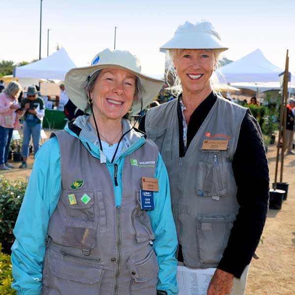 Two volunteers wearing vests and hats smile at an outdoor event with tents visible in the background.