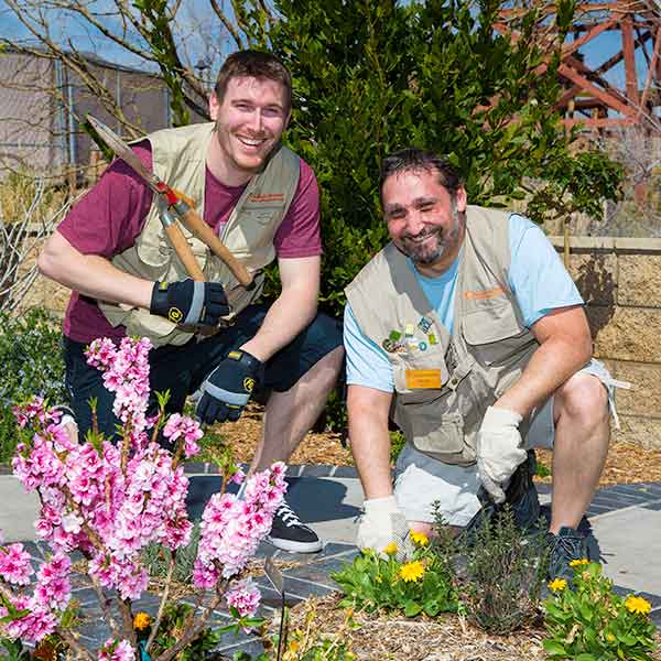 Two volunteers wearing aprons and gloves are kneeling in a garden full of blooming flowers, one holding a shovel, smiling at the camera.
