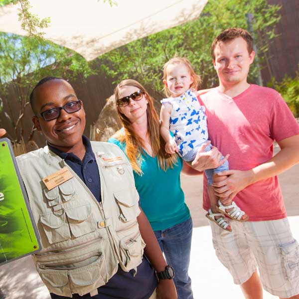 A volunteer standing with a family of three, including a toddler being held, at an outdoor exhibit.